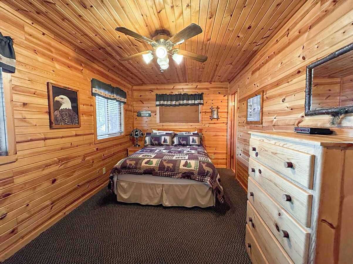 A wooden cabin bedroom with a bed, dresser, ceiling fan, and a framed picture of an eagle.