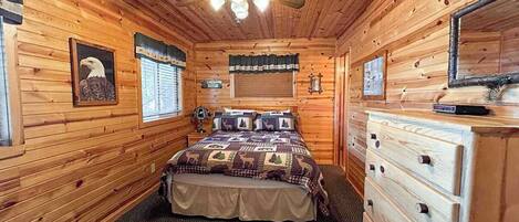 A wooden cabin bedroom with a bed, dresser, ceiling fan, and a framed picture of an eagle.