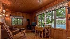 Interior de una cabaña con paredes de madera, chimenea, televisor, mecedora y ventanas amplias con vista a un lago.