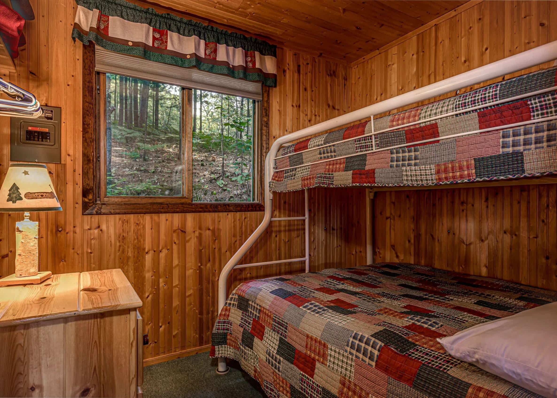 A wooden cabin room with a bunk bed, a small desk with a lamp, and a window overlooking a forest.