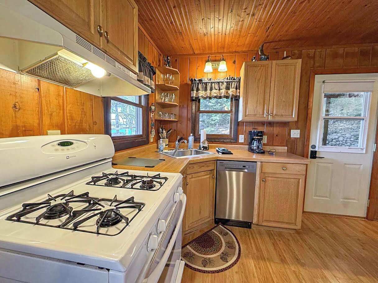 Une cuisine avec des meubles de rangement en bois, une cuisinière blanche, un lave-vaisselle et une fenêtre avec des rideaux.