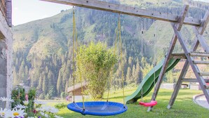 Un columpio de madera con un trampolín y un tobogán en un césped con montañas al fondo.