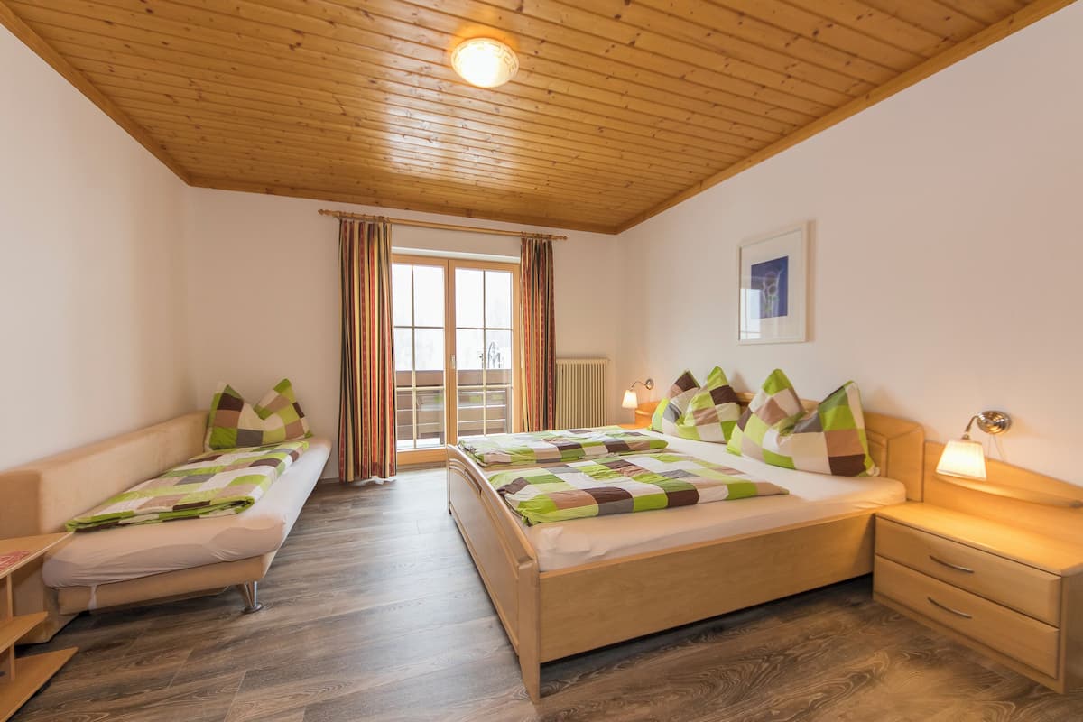 Ein Zimmer mit Holzboden, einem Bett mit Steppdecke, einem Nachttisch mit Lampe und einem Fenster mit Vorhängen.