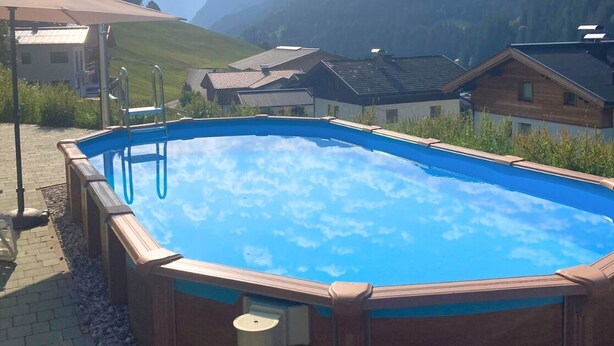 Una piscina con plataforma de madera, rodeada de casas y montañas.