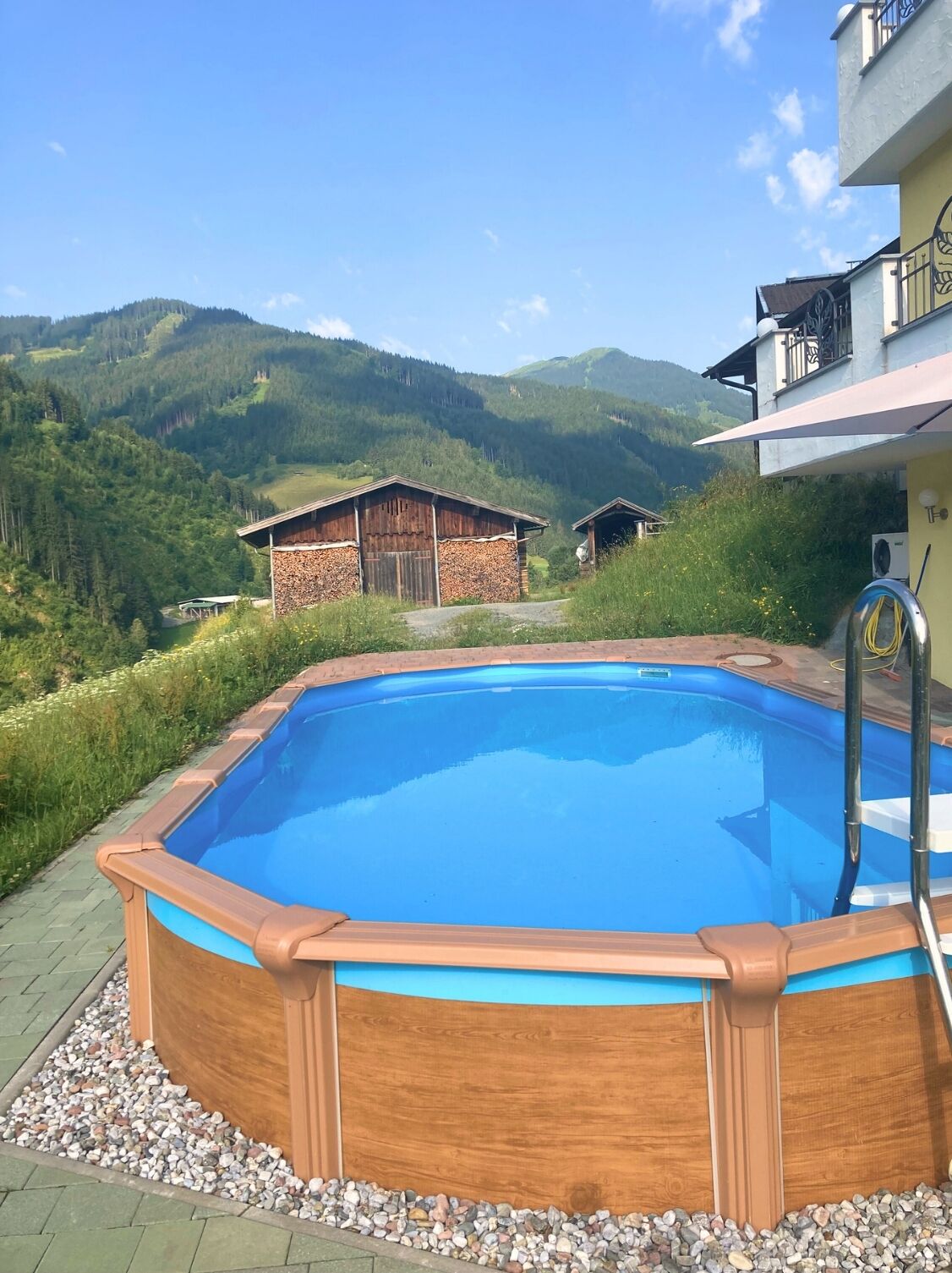 A swimming pool with a wooden deck, surrounded by a pebble border, with a view of mountains and a building in the background.