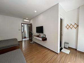 Una habitación de hotel con cama, escritorio con televisor, ventilador y estante con objetos.