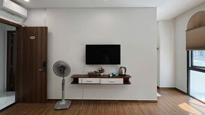 Una habitación moderna con televisor montado, una puerta de madera con un número, un ventilador de pie y un pequeño estante con objetos.