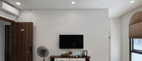 Une chambre moderne équipée d’un téléviseur fixé au mur, d’une porte en bois portant un numéro, d’un ventilateur sur pied et d’une petite étagère avec quelques objets.
