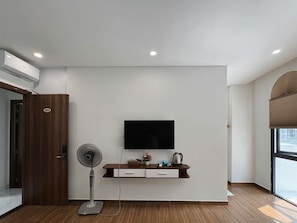 Une chambre moderne équipée d’un téléviseur fixé au mur, d’une porte en bois portant un numéro, d’un ventilateur sur pied et d’une petite étagère avec quelques objets.