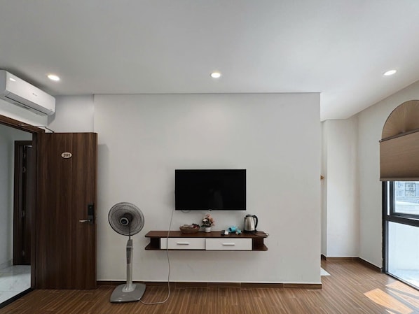 Una habitación moderna con televisor montado, una puerta de madera con un número, un ventilador de pie y un pequeño estante con objetos.