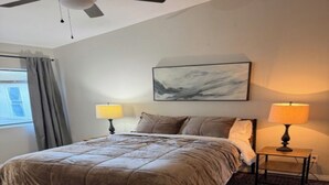 A bedroom with a bed, two bedside lamps, a nightstand, a wooden table, a ceiling fan, a window with curtains, and a painting on the wall. - SUMMER TIME SPECIALS!
Enjoyable 2-bedroom condo with AC in vibrant Tempe. (Tempe)