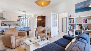 Uma sala moderna com uma mesa de centro de vidro, poltronas de couro, um sofá escuro, uma estante e um espaço de jantar com toalha de mesa estampada.