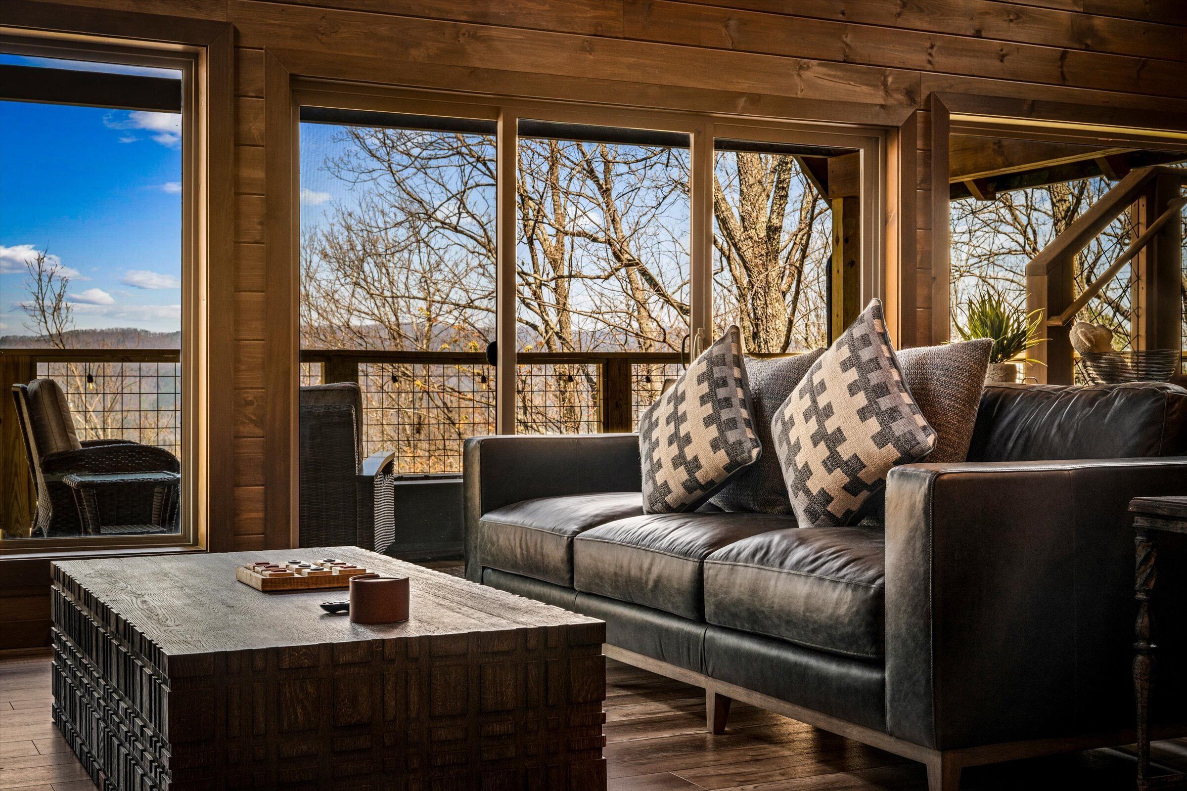 A modern living room with a leather sofa, wooden coffee table, and large windows offering a view of trees and hills.