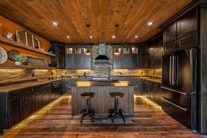 A modern kitchen with dark cabinetry, a central island with stools, a stone backsplash, and a wooden ceiling. - Kelly's Escape: A Rejuvenating Couple's Retreat (Sevierville)