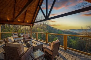 Una terraza de madera con mobiliario de mimbre, una fogata y vistas a un paisaje boscoso.