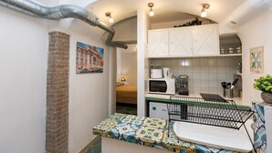 Een compacte keuken met een magnetron, broodrooster en spoelbak, een klein eethoekje met een tafel en stoelen, en een bakstenen muur met een schilderij.
