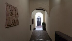 Een hal met stenen vloer, een witte muur met textuurrijk kunstwerk, een boogvormige doorgang en een donkere deur aan het einde.