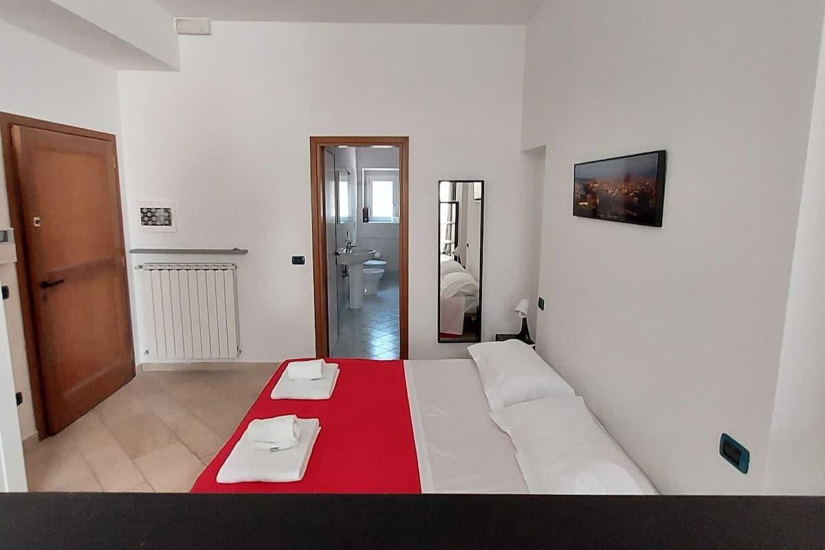 Habitación de hotel con cama, pared de acento roja, baño visible a través de una puerta abierta, un espejo y un cuadro en la pared.