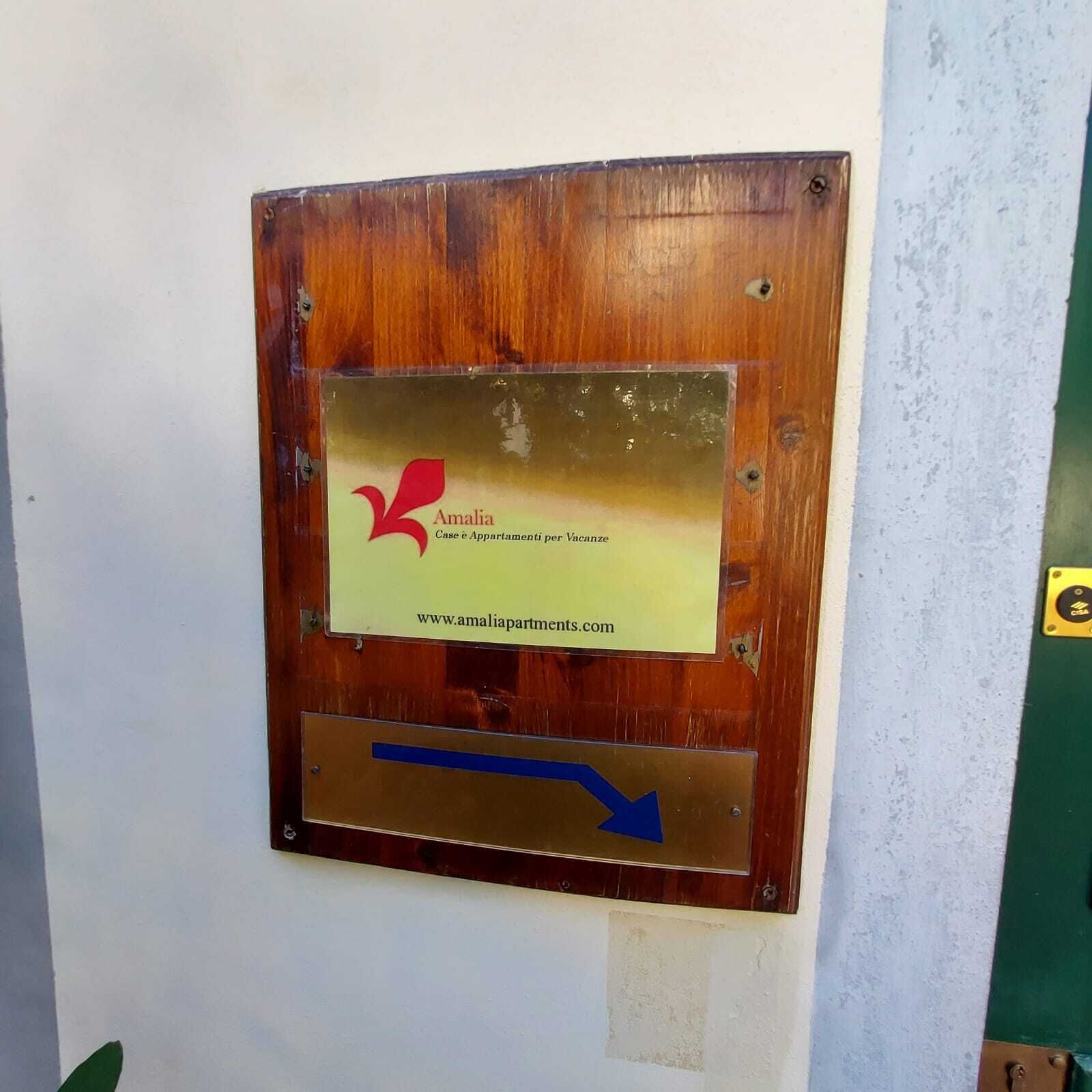 Una placa de madera con un logotipo y texto que indica 'Amalia Case è Appartamenti per Vacanze' y una dirección web.