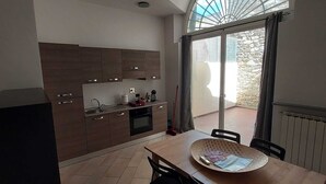 Een compacte keuken met moderne kastjes, een eethoek met een grote tafel en stoelen, en uitzicht door een glazen deur op een terras.