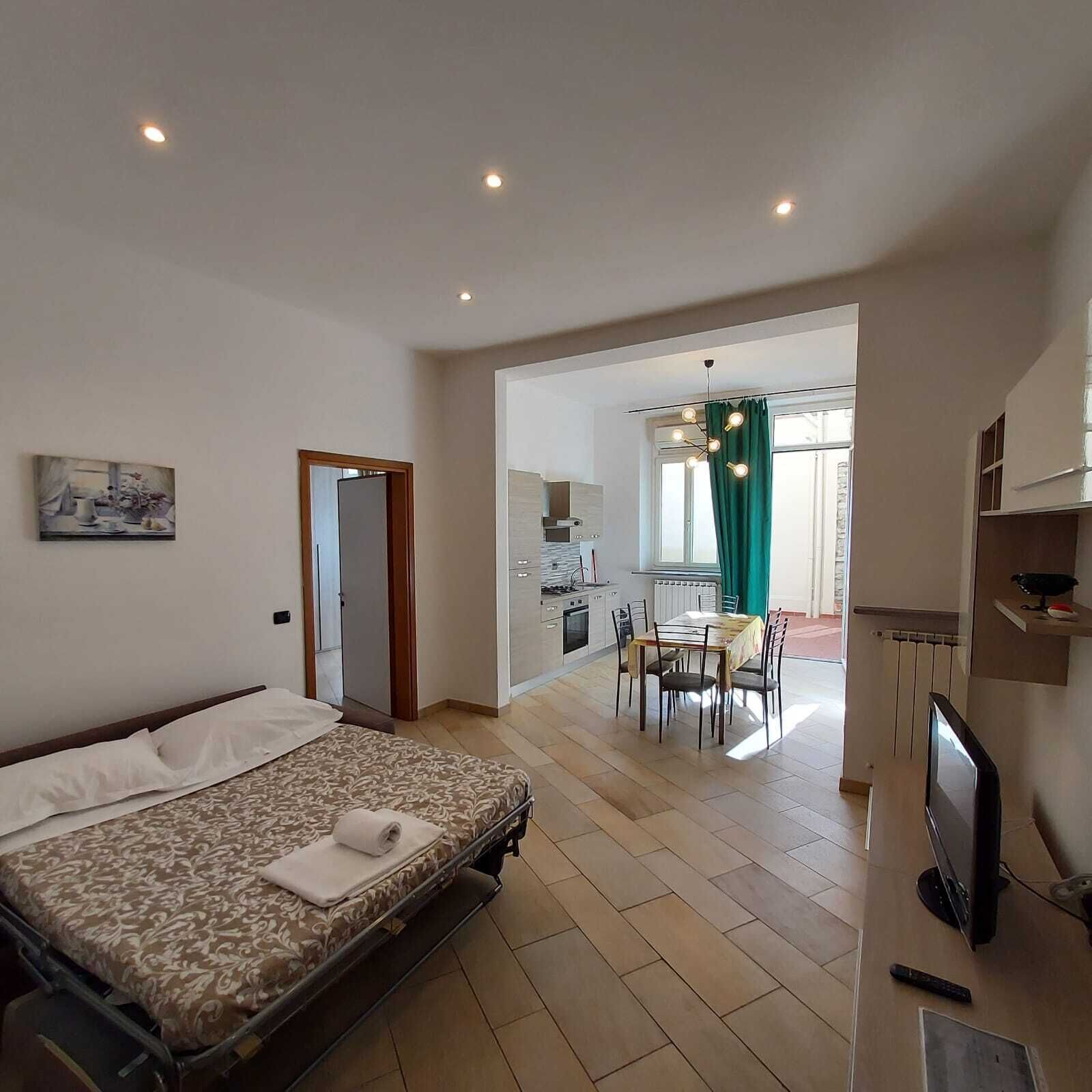 Ein Hotelzimmer mit einem Bett, einem Schreibtisch mit Fernseher, einer Essecke mit Tisch und Stühlen sowie einer durch eine offene Tür sichtbaren Küche.