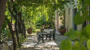 Un'area salotto all'aperto coperta, con tavoli e sedie, immersa nel verde e affiancata da un vialetto in pietra.