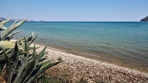 Ein freier Blick auf einen Kiesstrand, im Vordergrund Agavenpflanzen, ruhiges Meer und entfernte Landformen.