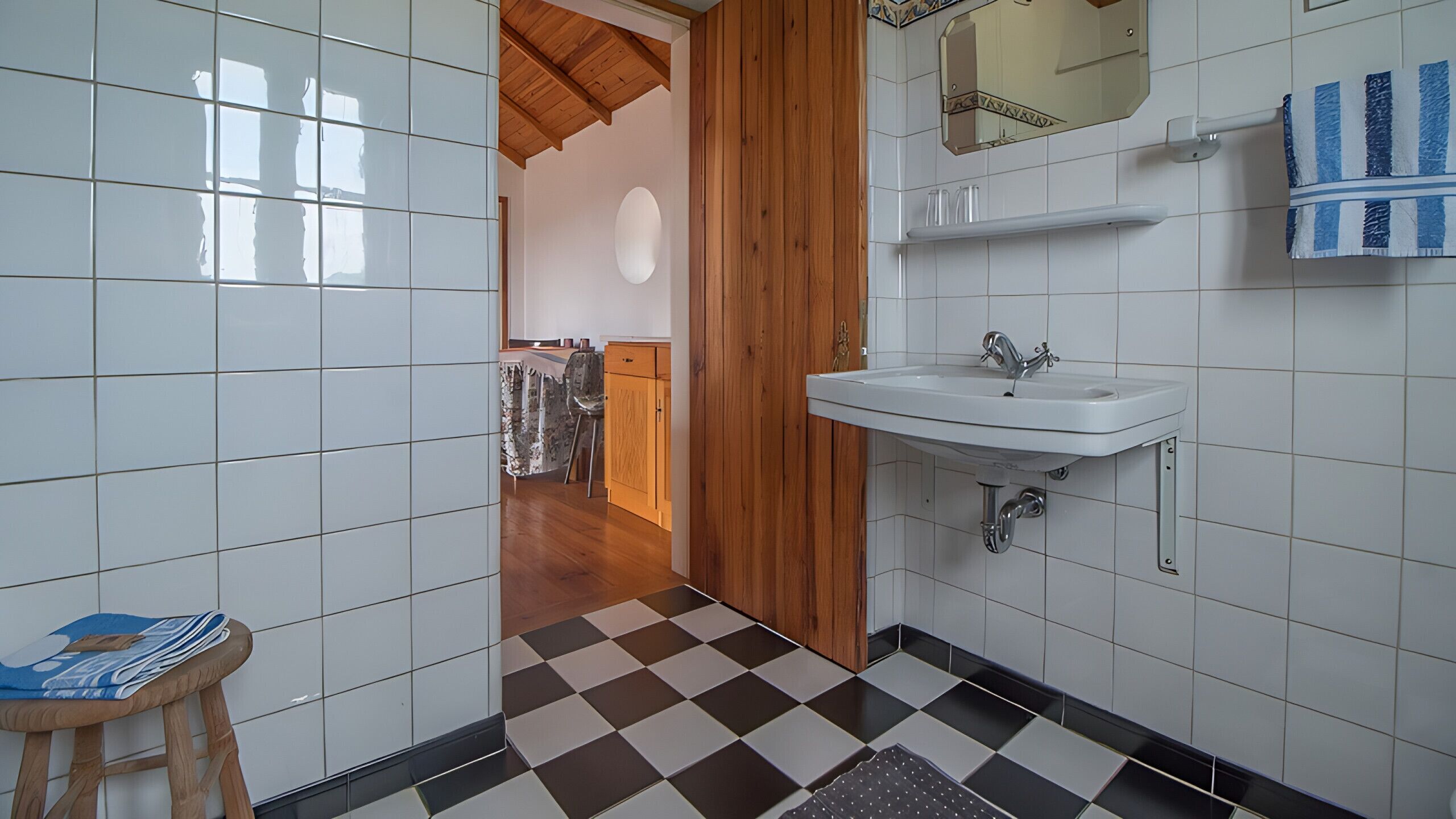 A bathroom with white tiles, a checkered floor, a wooden door, a sink, and a mirror.