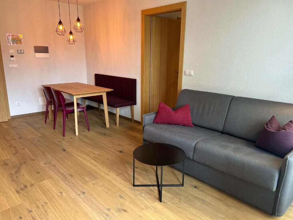 Ein Wohnzimmer mit grauem Sofa, einem Holzesstisch mit Stühlen, einem kleinen schwarzen Beistelltisch und Pendelleuchten.