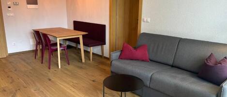 Ein Wohnzimmer mit grauem Sofa, einem Holzesstisch mit Stühlen, einem kleinen schwarzen Beistelltisch und Pendelleuchten.