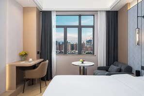 Une chambre d’hôtel équipée d’un lit, d’un bureau, d’une chaise, d’une petite table ronde et offrant une vue sur le paysage urbain grâce à de grandes fenêtres.
