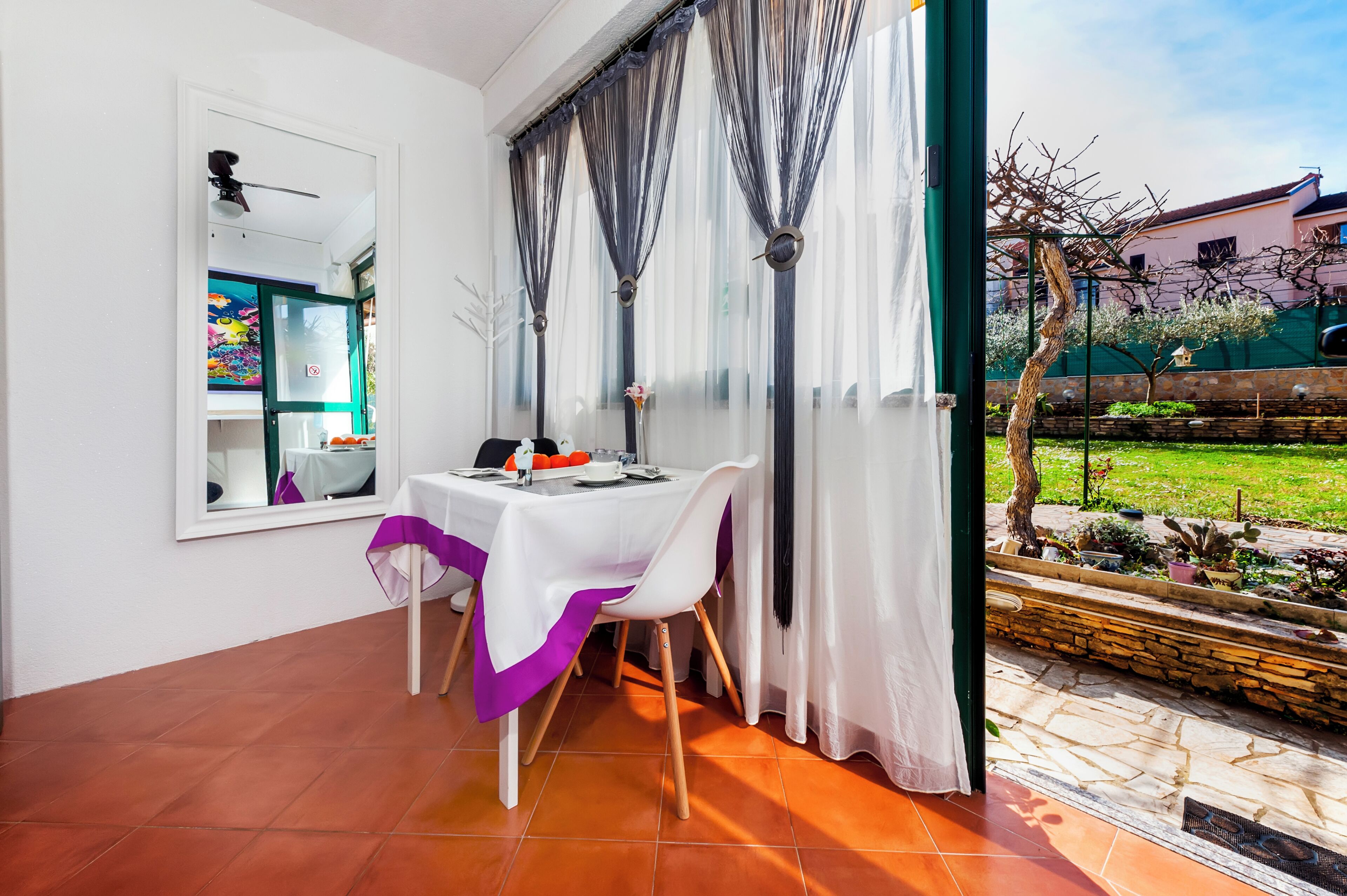 Un coin repas avec une table dressée pour deux personnes, une fenêtre avec des rideaux et une vue sur un jardin.
