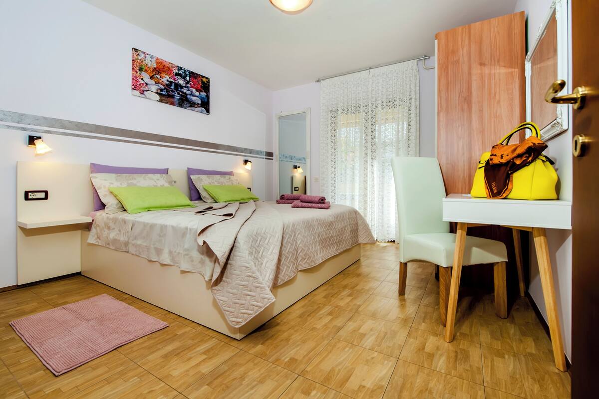 A bedroom with a bed, bedside tables, a desk, a chair, a mirror, and a door.