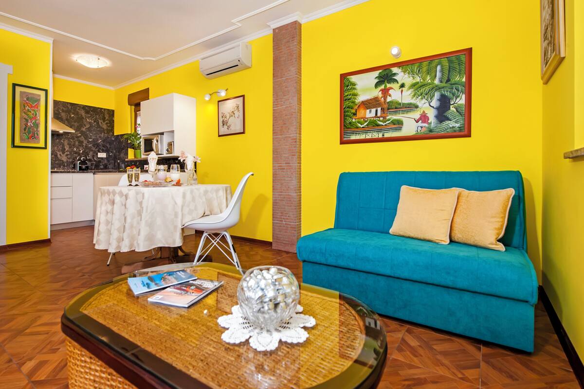 A bright yellow room with a blue sofa, a dining table with a white tablecloth, and a kitchen area.