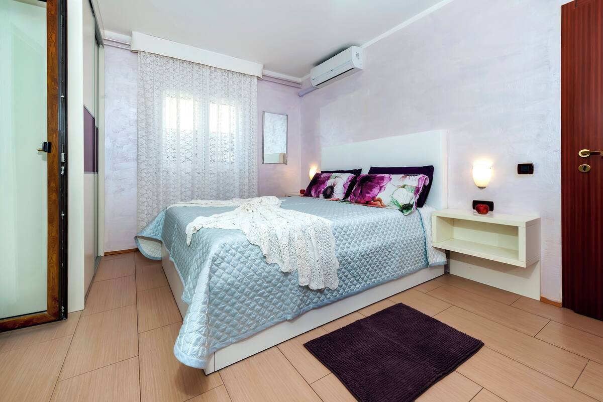 A bedroom with a bed, a bedside table, a nightstand, a wall-mounted air conditioner, and a door.