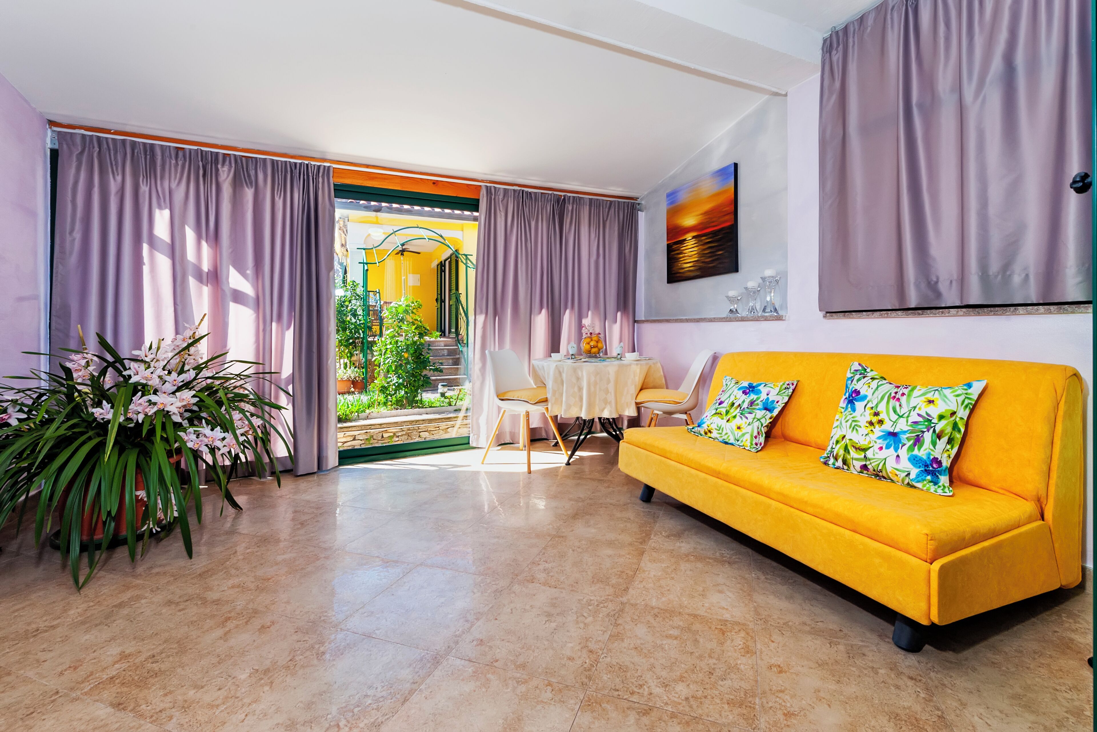 Un sofá amarillo brillante con cojines estampados, una mesa blanca con sillas y una planta grande en maceta.
