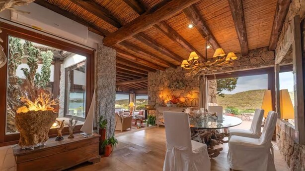 Una habitación amplia con techo de madera, una mesa redonda de vidrio, sillas blancas y una chimenea de piedra.