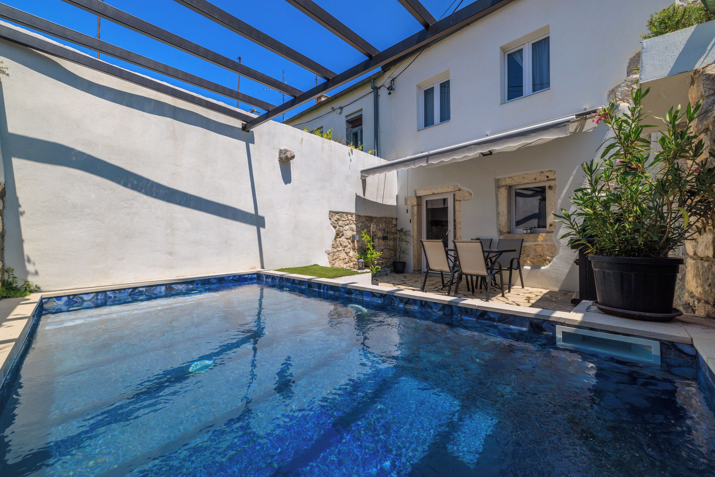 A swimming pool with a stone border, a patio with chairs, and a building with a white exterior and windows.
