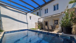Uma piscina com borda de pedra, um pátio com cadeiras e um edifício com fachada branca e janelas.