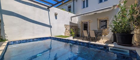 A swimming pool with a stone border, a patio with chairs, and a building with a white exterior and windows.
