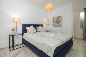 Une chambre à coucher comprenant un lit, deux tables de chevet avec des lampes, un luminaire au plafond, une œuvre d’art murale et une fenêtre avec des rideaux.