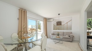 Una sala de estar moderna con una mesa de comedor de vidrio, sillas blancas, un sofá y una lámpara colgante.