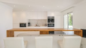 Una cocina moderna con armarios blancos, encimera de madera y horno empotrado. Hay tres sillas blancas frente a la encimera.