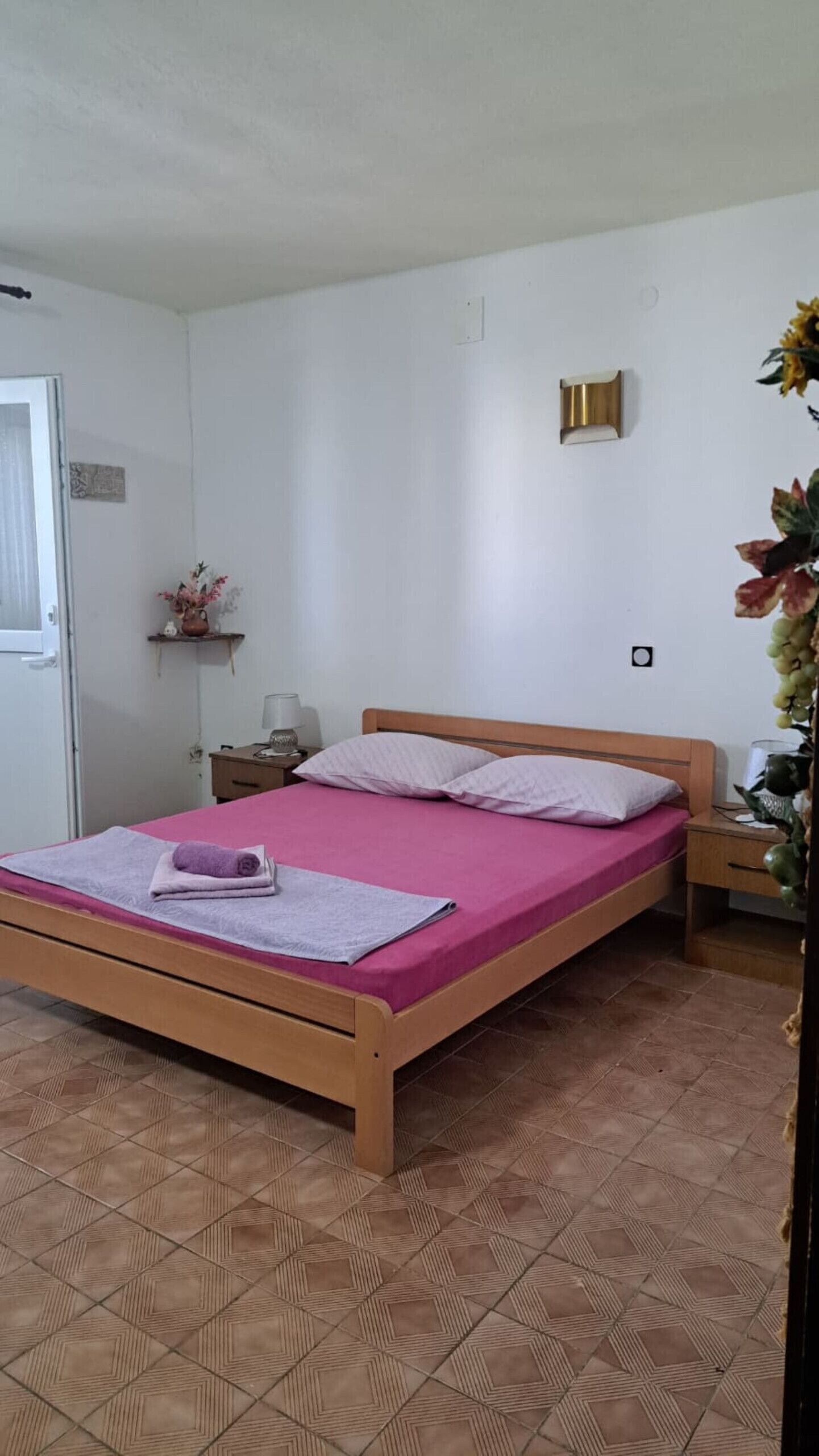 Habitación con cama individual de estructura de madera, colcha rosa, mesita de noche pequeña con lámpara y lámpara de pared.