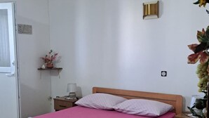 Une chambre avec un lit simple, un cadre en bois, une couvre-lit rose, une petite table de chevet avec une lampe et un luminaire fixé au mur.