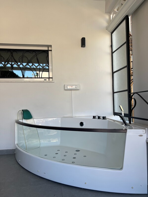 A modern bathroom with a glass-enclosed bathtub, a window with a decorative metal frame, and a control panel on the wall. - Casa Divinus (Athens)