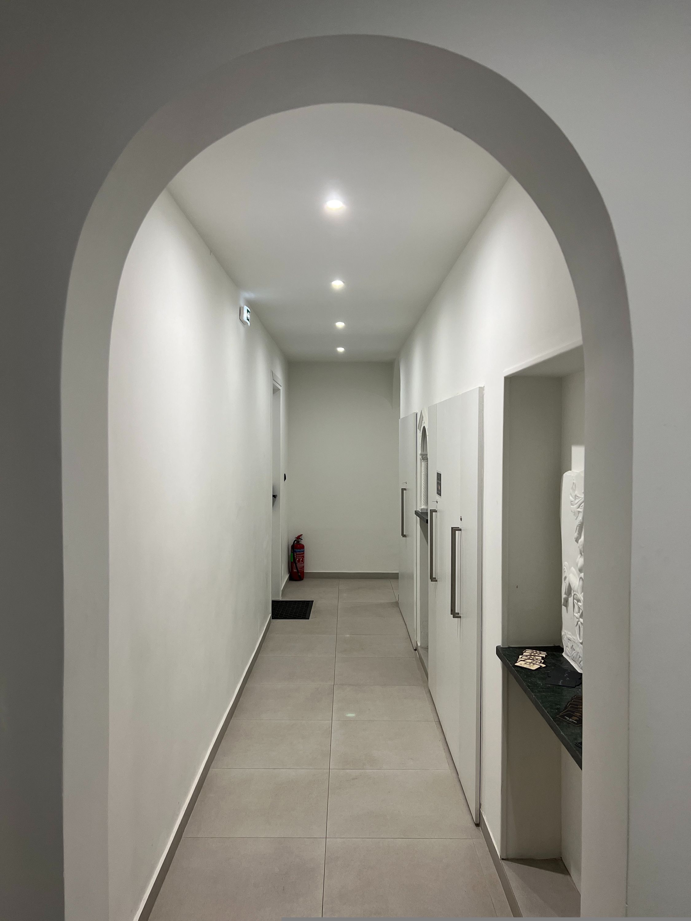 A hallway with an arched ceiling, recessed lighting, a fire extinguisher, and a tiled floor.