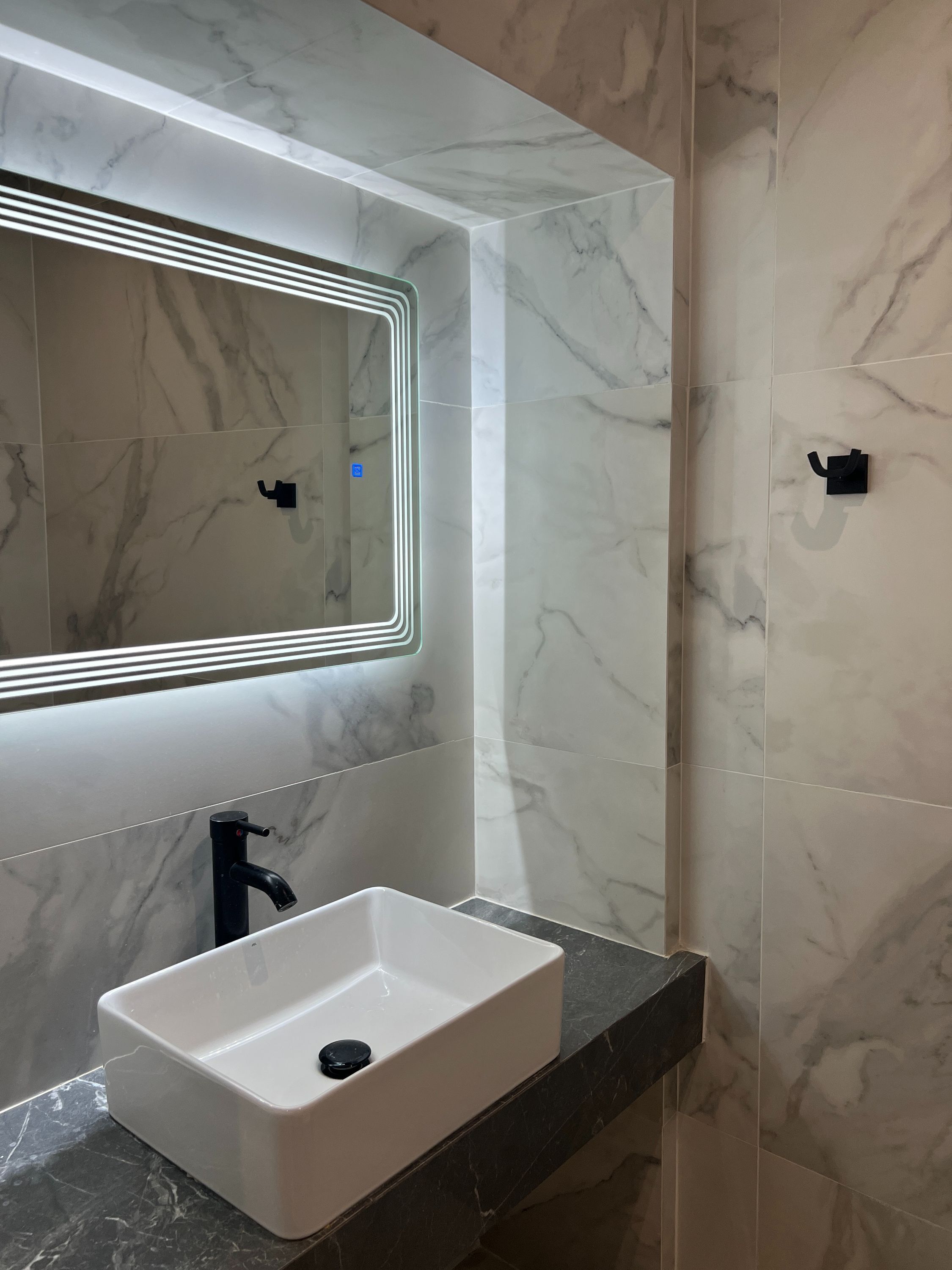 Een badkamer met een wit wastafel, een zwarte kraan, een spiegel met LED-verlichting en wanden met een marmerprint.