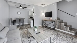 A modern living room with a grey sofa, a coffee table with a plant, a TV on a stand, and a staircase. - Cozy 1BR townhome perfect for relaxing stays (Arlington)