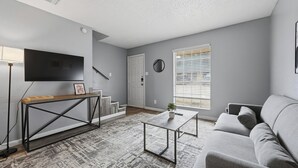 A modern living room with a grey sofa, a coffee table, a TV on the wall, and a staircase leading to another level. - Cozy 1BR townhome perfect for relaxing stays (Arlington)